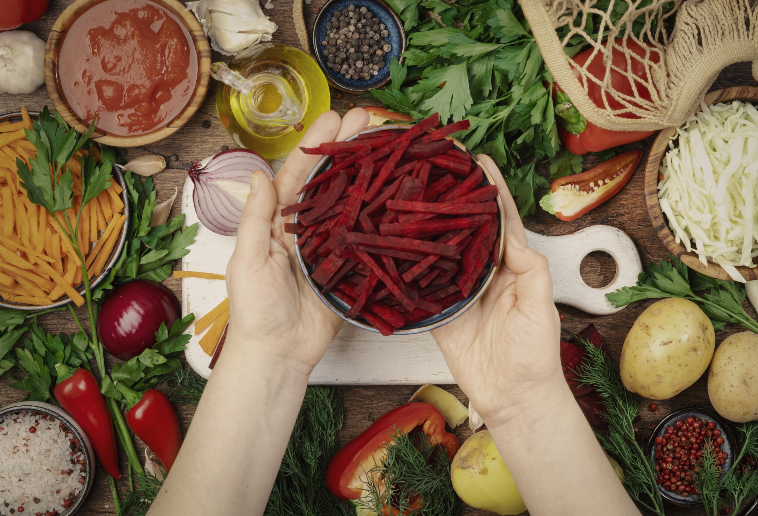 Fresh vegan vegetarian ingredients, vegetables, spices, herbs for healthy eating on rustic wooden kitchen table with cutting board. Womens hands holding chopped beets. Diet, clean, comfort food cooking concept